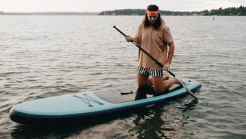 Man with long hair and Orange headband kneeling on blue paddleboard whilst out on the sea.