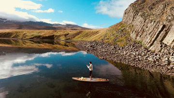 Person kayaking in open water surrounded by mountains and a grass covered landscape